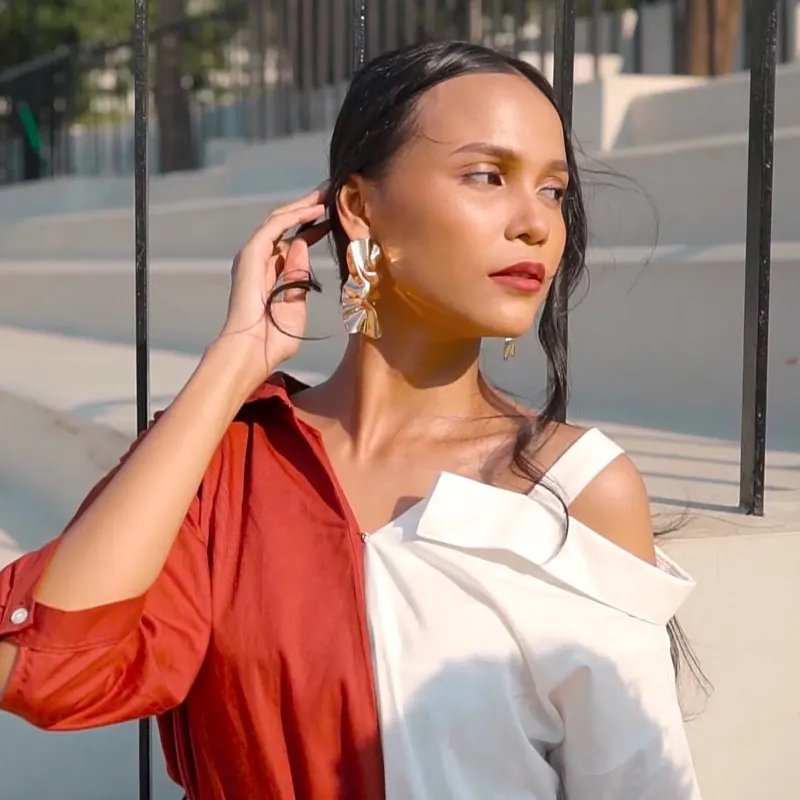 Model berkulit sawo matang dengan kemeja merah bata dan putih off-shoulder merapikan rambut, cahaya matahari sore